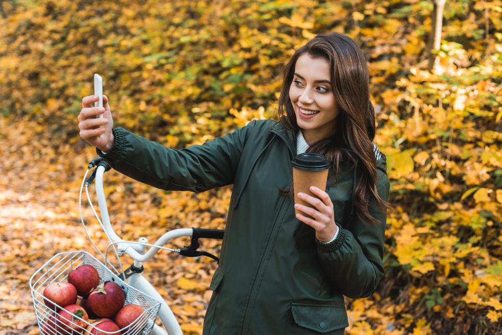 selfie 秋の森でのスマート フォンの使い捨てのコーヒー カップの陽気な美女  - 写真・画像