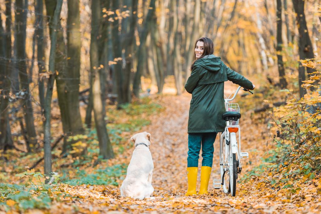 smiling woman with bicycle and her golden retriever sitting near on yellow leafy path in forest  - Photo, Image