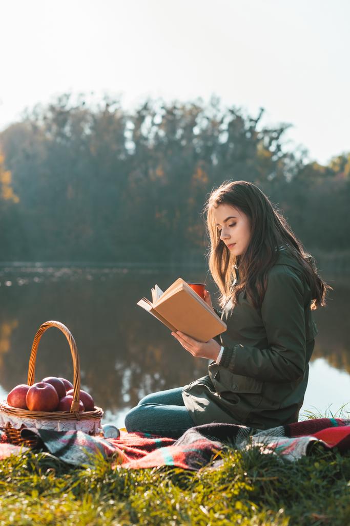 Widok z boku z atrakcyjna młoda kobieta z filiżanki kawy, czytanie książki na koc w pobliżu staw w parku - Zdjęcie, obraz