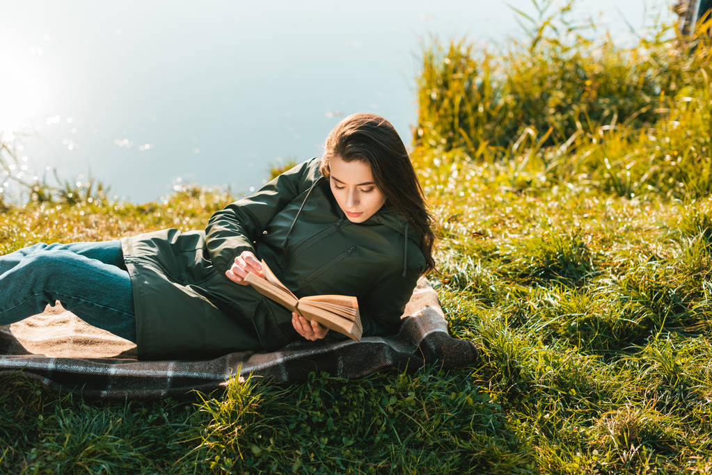 όμορφη γυναίκα διαβάζοντας το βιβλίο βάζοντας την κουβέρτα κοντά Λίμνη στο πάρκο - Φωτογραφία, εικόνα