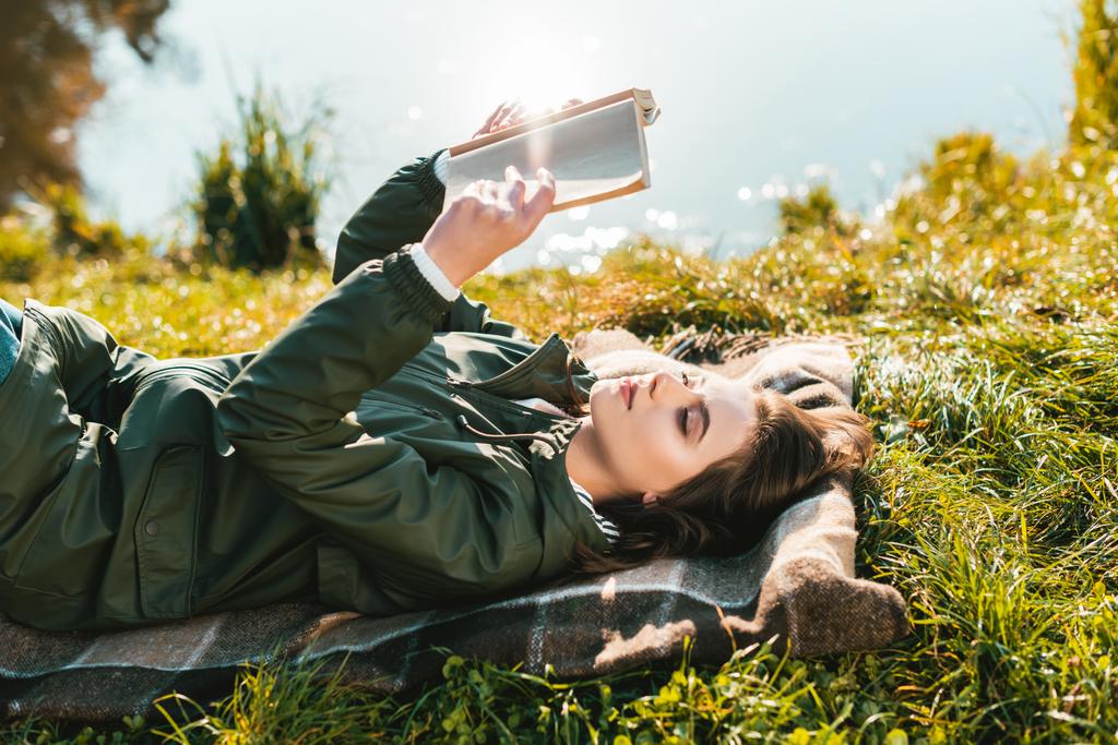 side view of cheerful young woman reading book while laying on blanket near pond in park - Photo, Image
