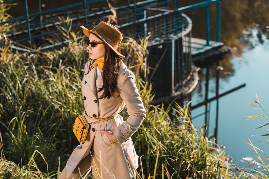 приваблива молода жінка в сонцезахисних окулярах, тренч пальто і капелюх позує біля ставка в парку
 - Фото, зображення