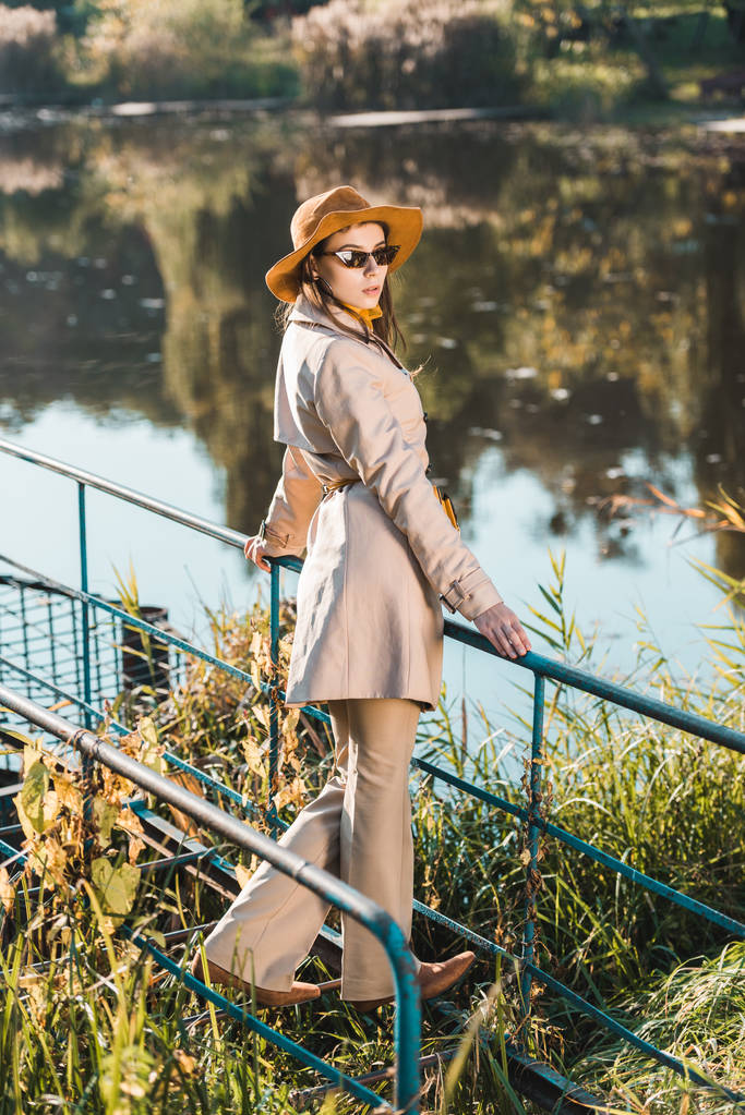 селективный фокус стильной женской модели в солнцезащитных очках, плаще и шляпе, позирующих возле пруда в парке
 - Фото, изображение