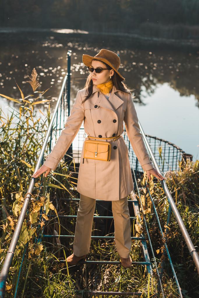 mladá žena módní sluneční brýle, plášť a klobouk pózuje u rybníka v parku - Fotografie, Obrázek