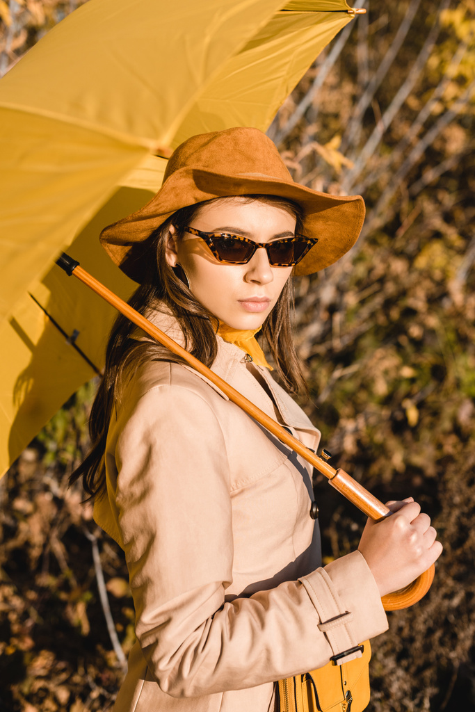 stylové elegantní žena v sluneční brýle a klobouk při pohledu na fotoaparát a držení žlutý deštník venku  - Fotografie, Obrázek
