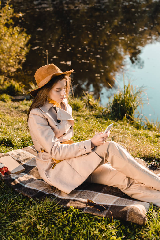 élégante jeune femme en chapeau en utilisant smartphone tout en étant assis sur la couverture près de l'étang dans le parc
 - Photo, image