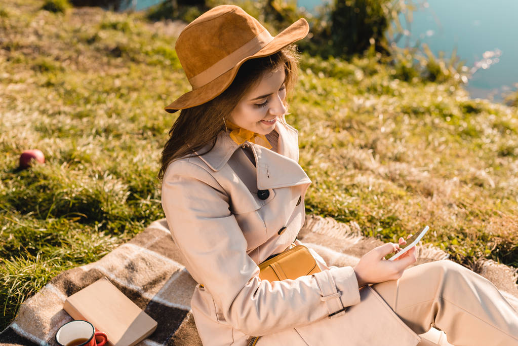 visão de alto ângulo de mulher elegante sorridente em chapéu usando smartphone enquanto sentado em cobertor ao ar livre
  - Foto, Imagem