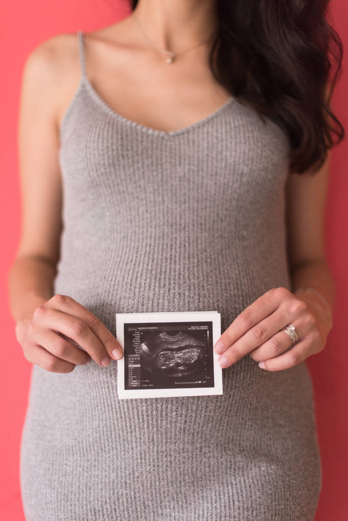усміхнена вагітна жінка показує ультразвукове зображення її ненародженої дитини ізольовано на червоному тлі
 - Фото, зображення