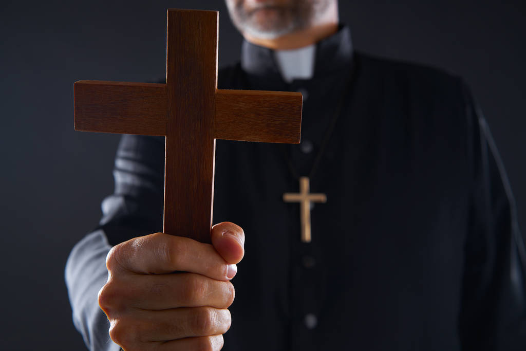 Sacerdote segurando cruz de madeira orando em primeiro plano
 - Foto, Imagem