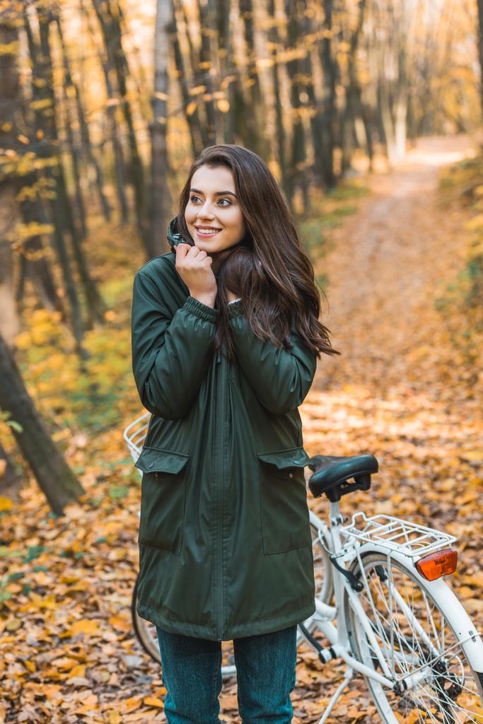 selectieve aandacht van lachende jonge vrouw weg op zoek in de buurt van fiets in herfst bos - Foto, afbeelding