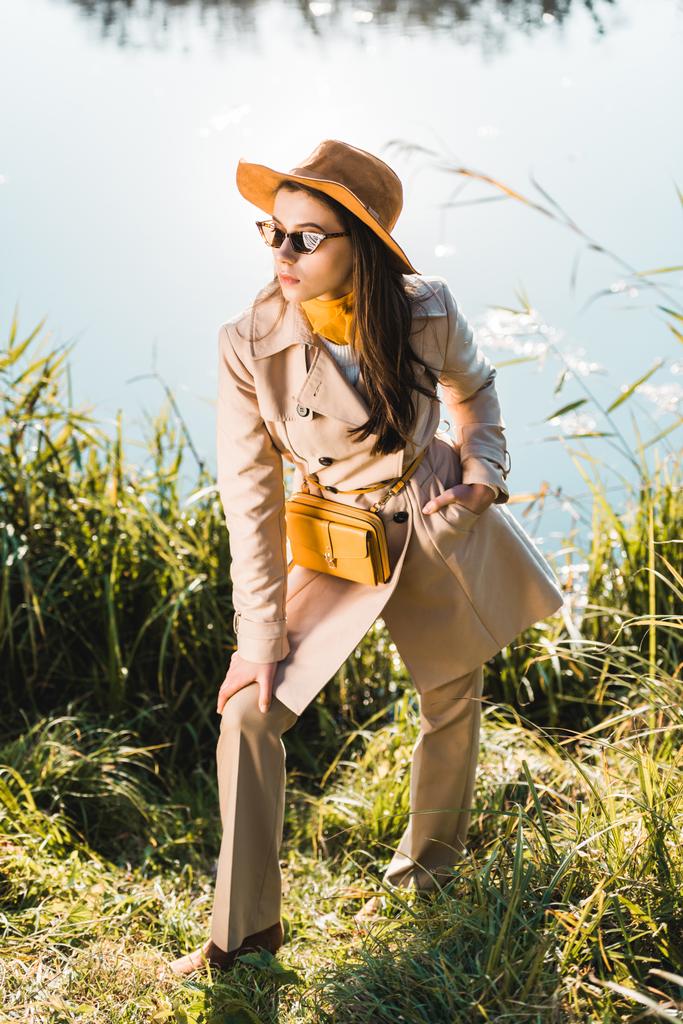 élégante femme élégante en trench coat, chapeau et lunettes de soleil posant près de l'étang à l'extérieur
 - Photo, image