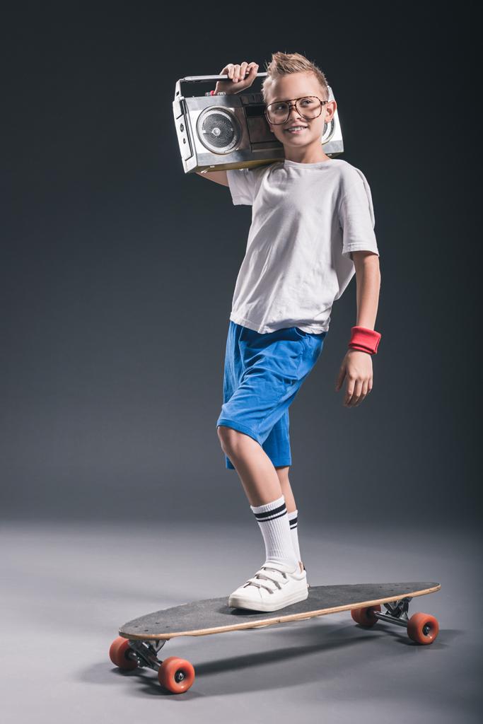 улыбающийся мальчик с бумбоксом на плече, стоящий на длинной доске на сером фоне
 - Фото, изображение