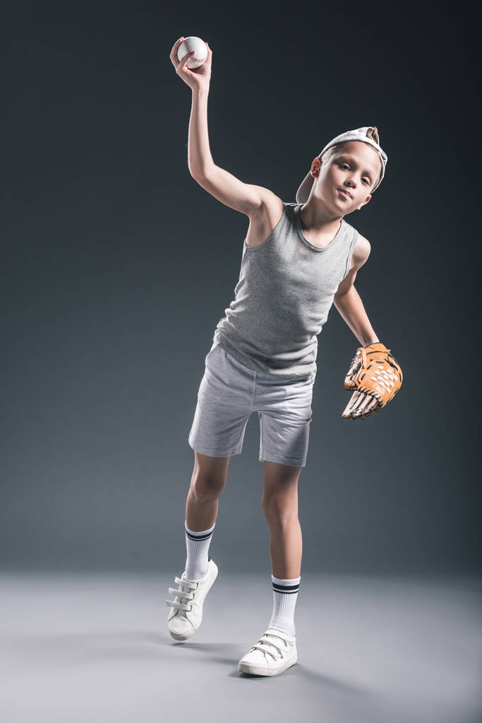 Bub in Mütze mit Baseballhandschuh wirft Ball vor grauem Hintergrund - Foto, Bild