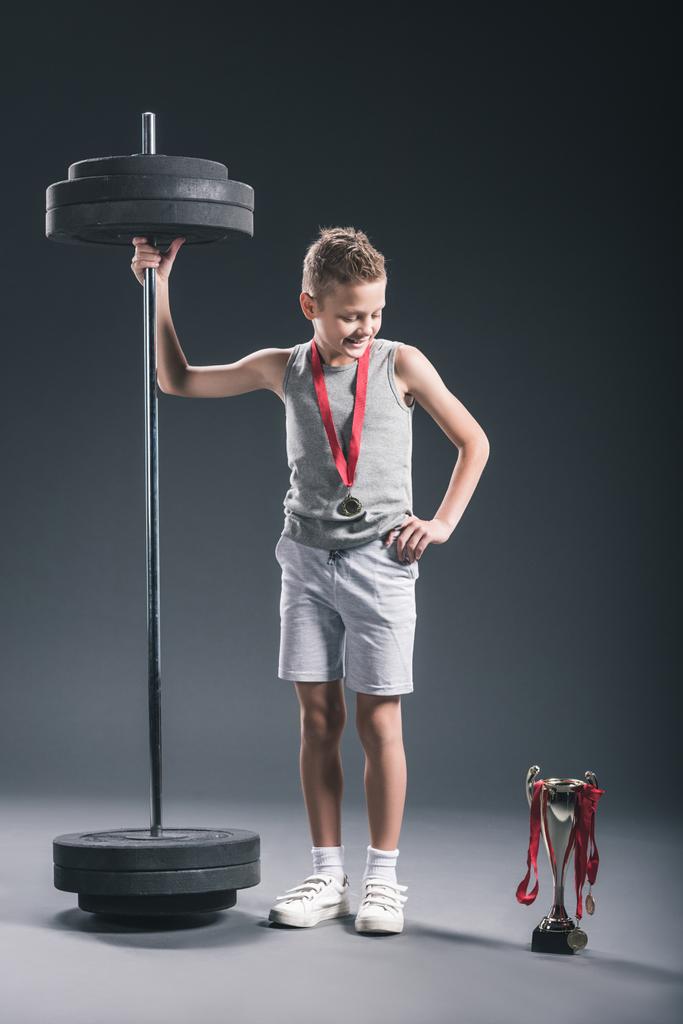 uśmiechnięty chłopiec w sportowej ze sztangą i medal, patrząc na Puchar Mistrzów na ciemnym tle - Zdjęcie, obraz