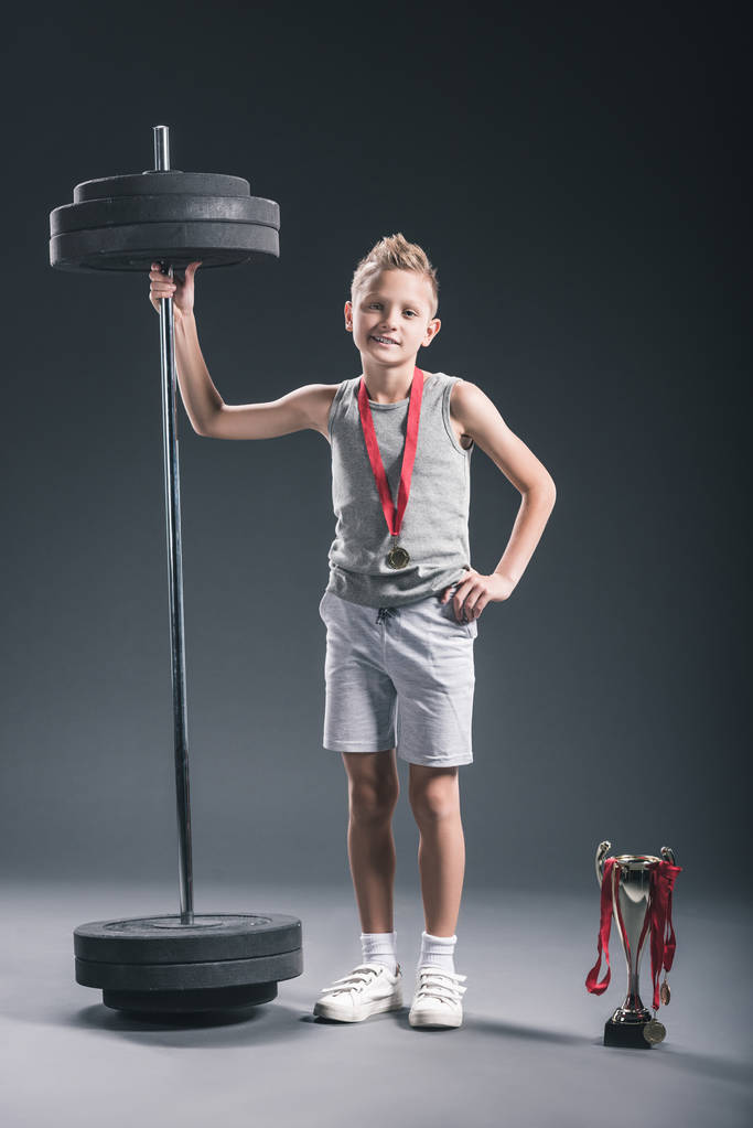 Vorpubertärer Junge in Sportbekleidung mit Langhantel, Meisterpokal und Medaillen vor dunklem Hintergrund   - Foto, Bild