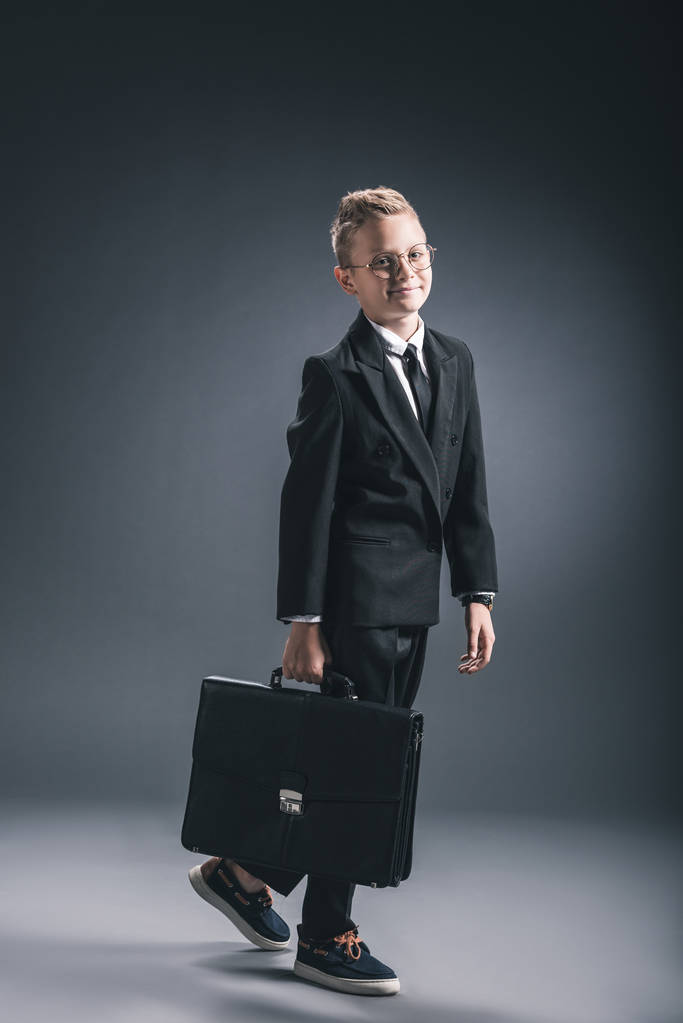 smiling boy in businessman suit and eyeglasses with suitcase on dark background - Photo, Image