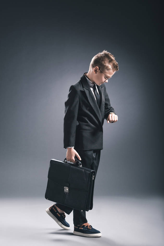 preteen αγόρι σε επιχειρηματία κοστούμι και γυαλιά με βαλίτσα ελέγχοντας φορά σε σκούρο φόντο - Φωτογραφία, εικόνα