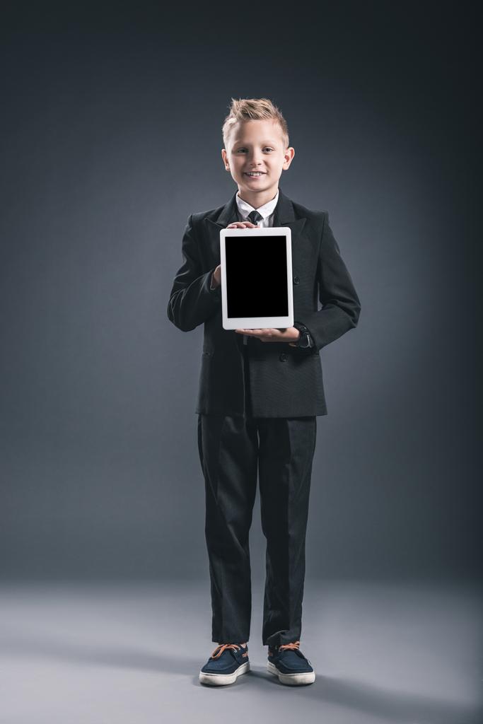 niño sonriente vestido como hombre de negocios mostrando tableta con pantalla en blanco en las manos sobre fondo gris
 - Foto, imagen