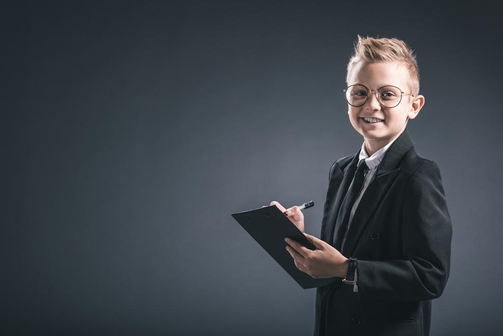 smiling preteen boy in businessman suit and eyeglasses with notepad on grey background - Photo, Image