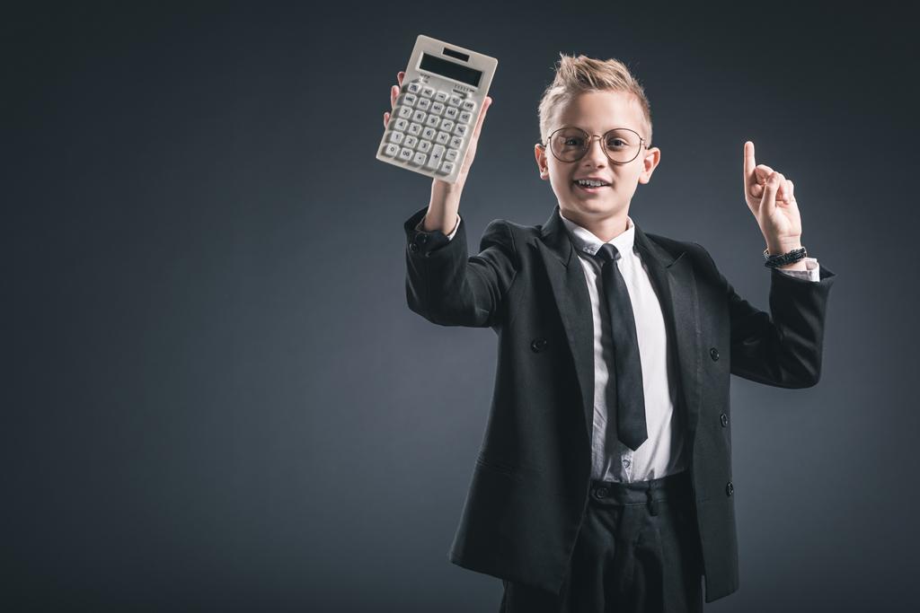 portrait of boy dressed as businessman in eyeglasses gesturing and showing calculator on dark background - Photo, Image