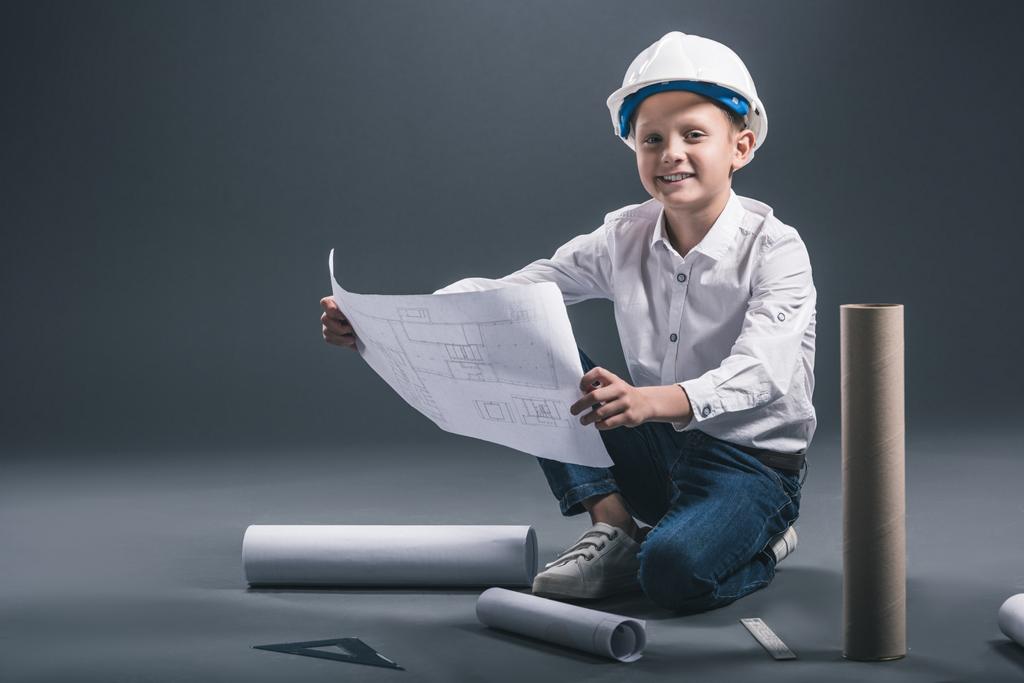 niño sonriente en casco de arquitecto sosteniendo planos sobre fondo oscuro
 - Foto, Imagen
