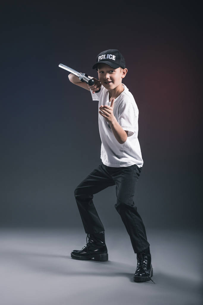 Мальчик-подросток в белой футболке и кепке полицейская форма с дубинкой позирует на темном фоне
 - Фото, изображение