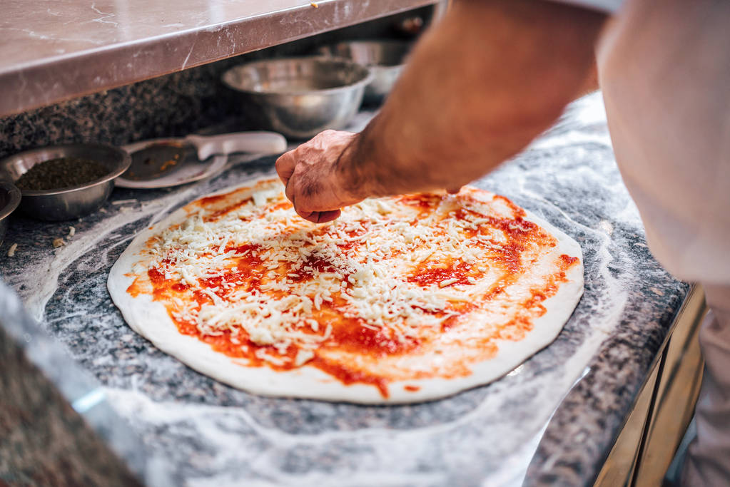 Der Koch bereitet die Pizza zu. Nahaufnahme. Käse auf Pizzaboden hinzufügen. - Foto, Bild