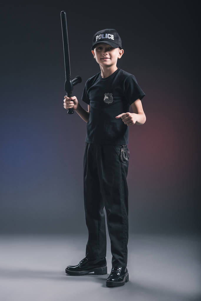 preteen boy in policeman uniform with truncheon on dark backdrop - Photo, Image