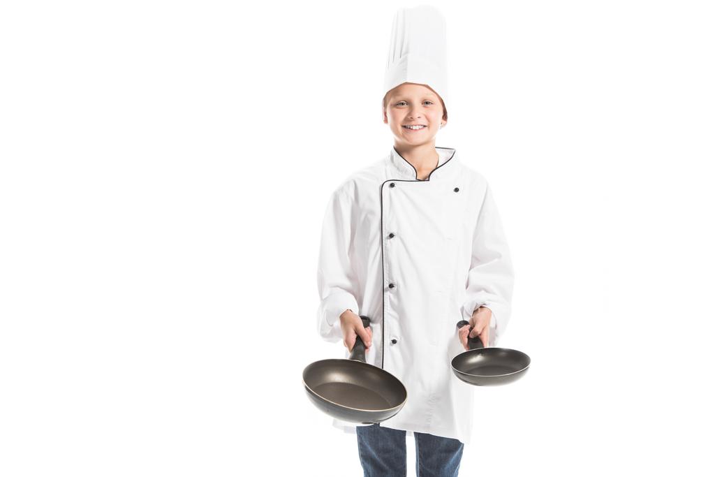 Lächelnder Junge in weißer Koch-Uniform und Hut, Bratpfannen haltend und isoliert auf weiß in die Kamera blickend - Foto, Bild