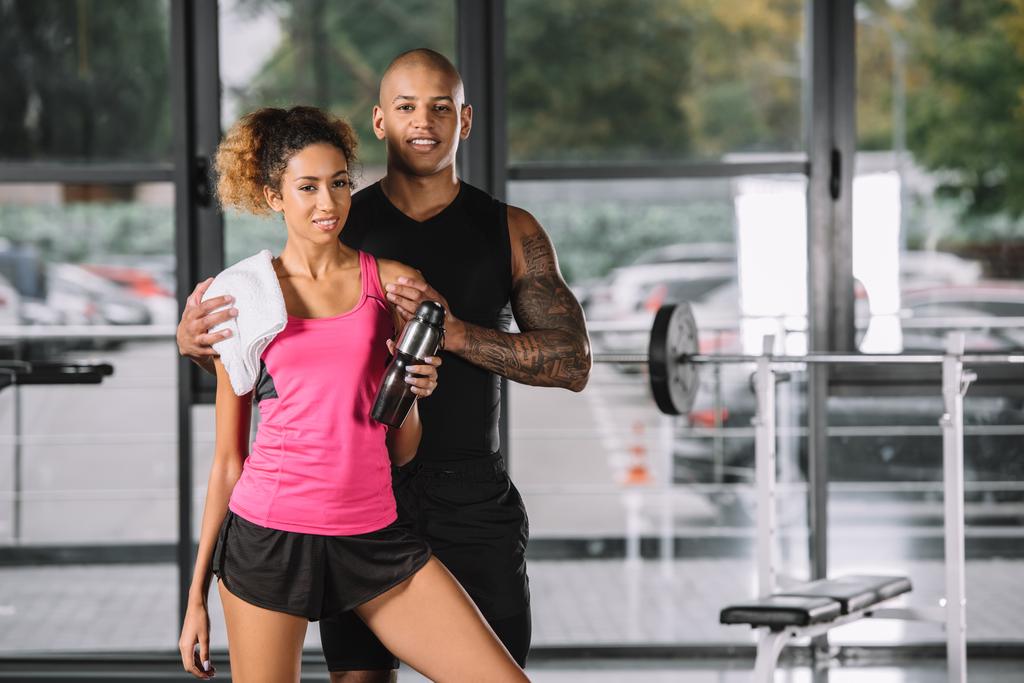 young african american sportsman embracing girlfriend with sport bottle and towel at gym - Photo, Image