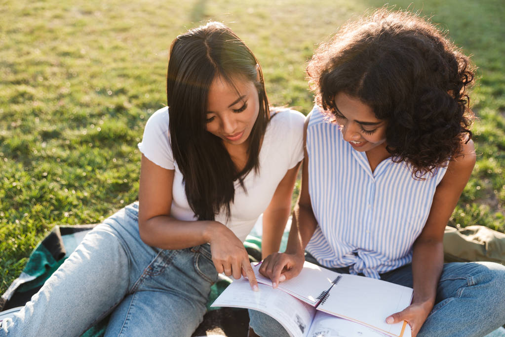 Two lovely young girls students sitting on a grass at the campus, studying with textbooks - Photo, Image