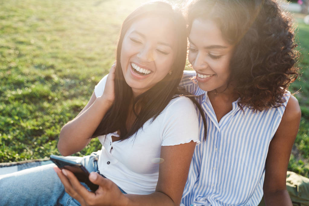 Изображение счастливых улыбающихся молодых друзей девушки в парке разговаривают друг с другом с помощью мобильного телефона
. - Фото, изображение