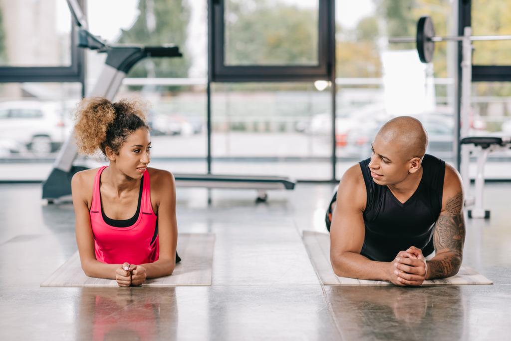 cheerful african american couple of athletes looking at each other while resting on fitness mats at gym - Photo, Image