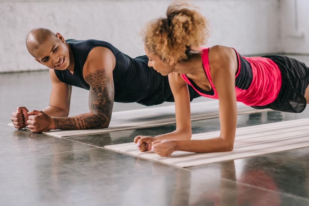 jovem casal de atletas afro-americanos olhando uns para os outros e fazendo prancha em tapetes de fitness no ginásio
 - Foto, Imagem