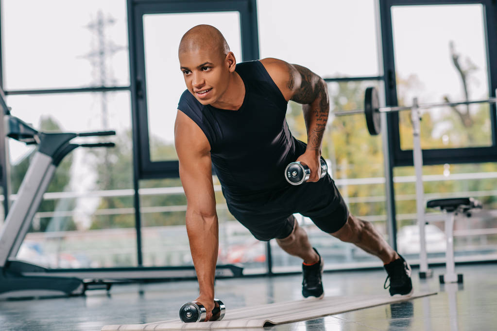 sorridente muscular afro-americano desportista exercitando com halteres no tapete de fitness no ginásio
 - Foto, Imagem