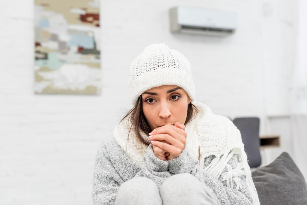 freezed young woman in warm clothes sitting on couch at home and blowing at hands - Photo, Image