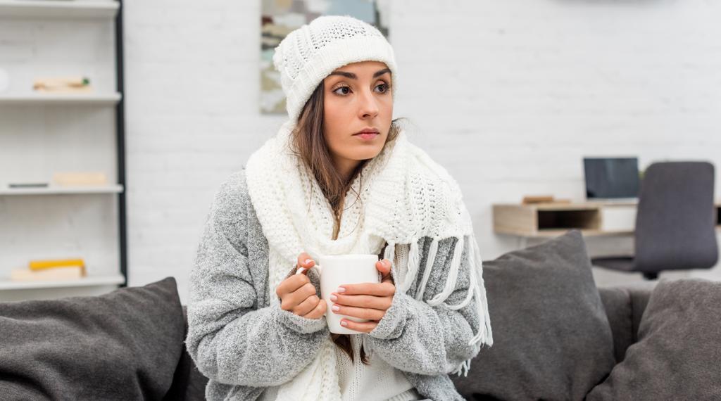 παγώνω νεαρή γυναίκα σε ζεστά ρούχα κρατώντας φλιτζάνι τσάι του πλανήτη ενώ κάθεται στον καναπέ στο σπίτι - Φωτογραφία, εικόνα