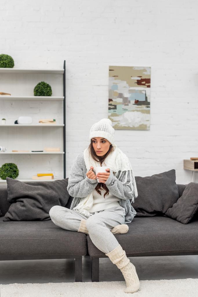 freezed young woman in warm clothes holding cup of hot tea while sitting on couch at home - Photo, Image