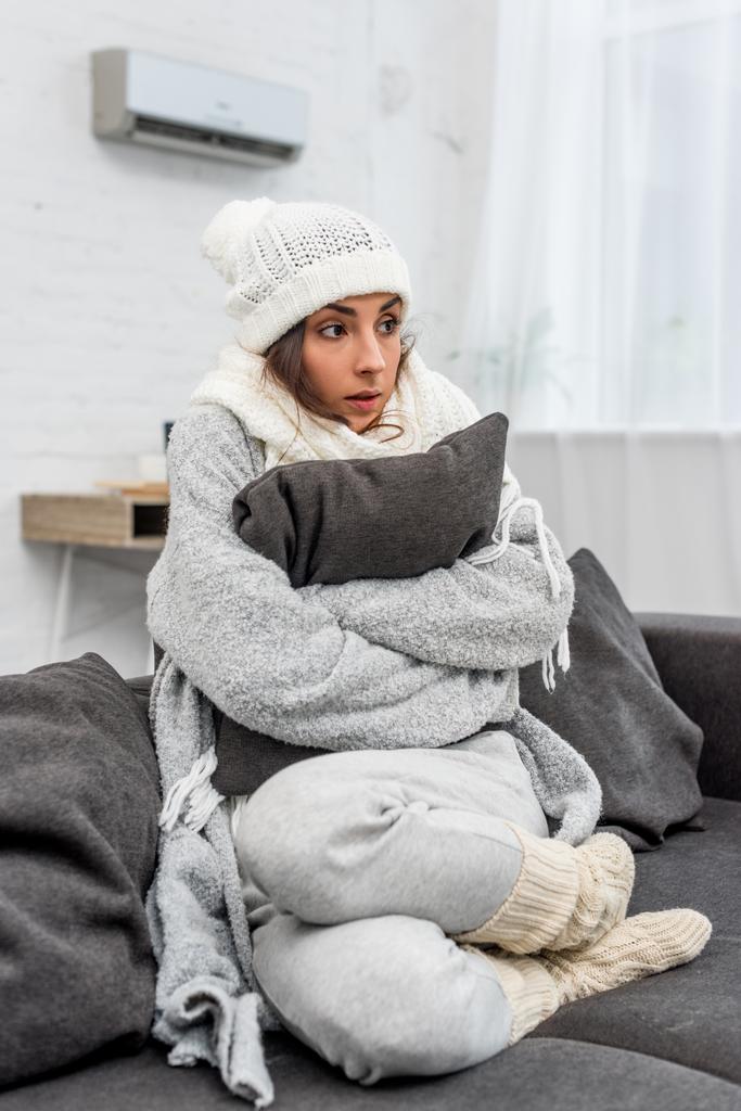 замерзшая шокированная женщина в теплой одежде сидит на диване и обнимает подушку дома
 - Фото, изображение