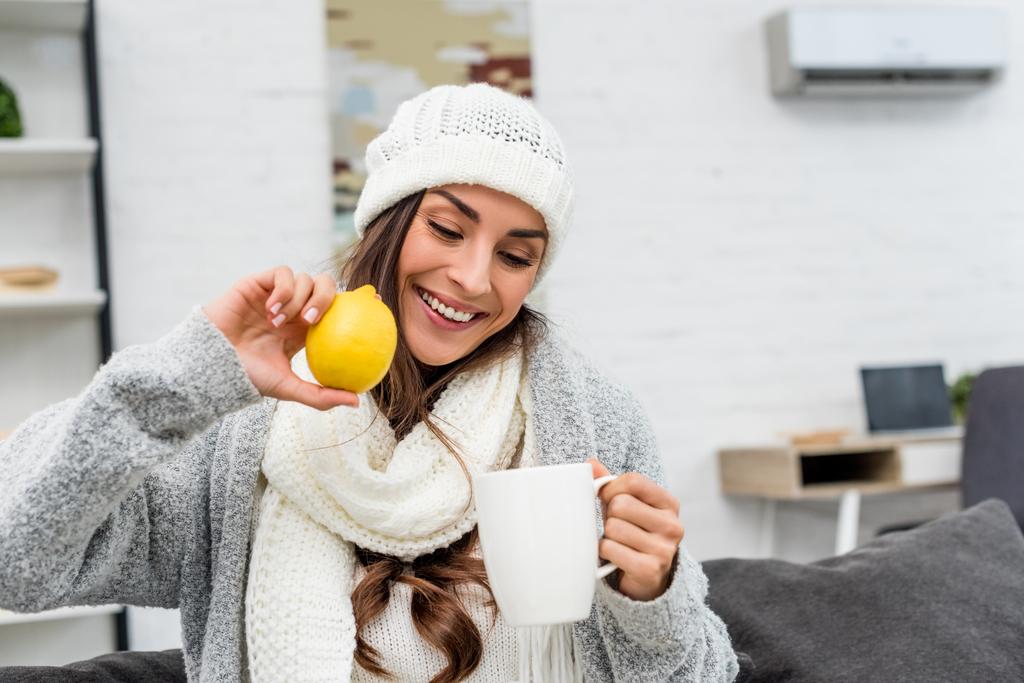 sonriente joven con ropa de abrigo sosteniendo limón y taza de té en casa
 - Foto, Imagen