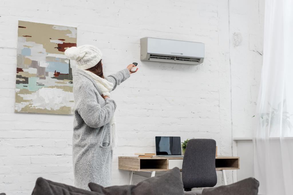beautiful young woman in warm clothes pointing at air conditioner with remote control at home - Photo, Image
