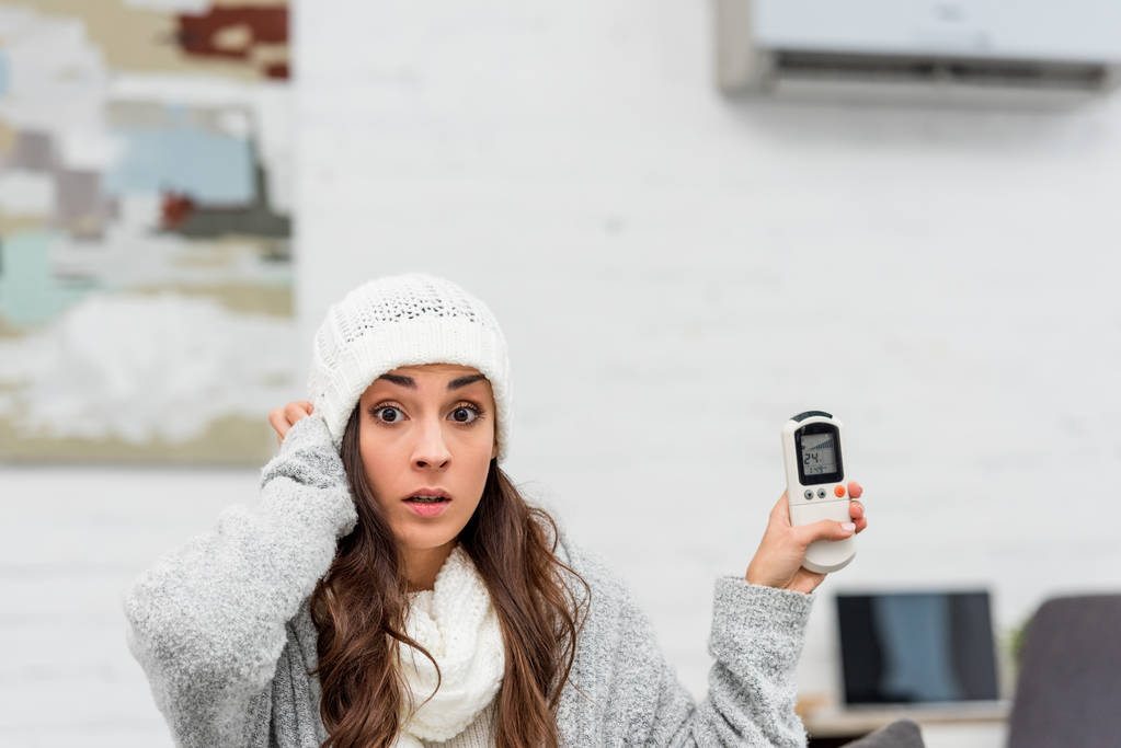 šokovaná mladá žena v teplé oblečení doma drží air conditioner dálkový ovladač - Fotografie, Obrázek