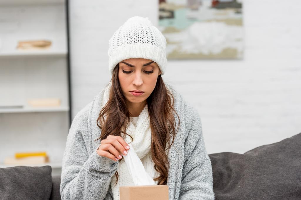 depressive, kranke Frau in warmer Kleidung nimmt Papierserviette aus Schachtel, während sie auf der Couch sitzt - Foto, Bild