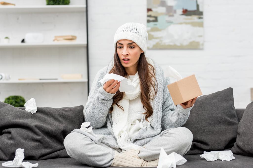 sick young woman in warm clothes sitting on messy couch and sneezing with paper napkins at home - Photo, Image