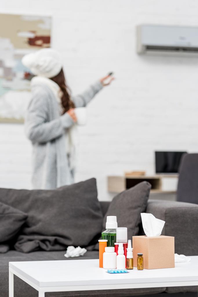 άρρωστη γυναίκα σε ζεστά ρούχα δείχνουν στο κλιματιστικό με το τηλεχειριστήριο με διάφορα φάρμακα που στέκεται στο τραπέζι σε πρώτο πλάνο - Φωτογραφία, εικόνα