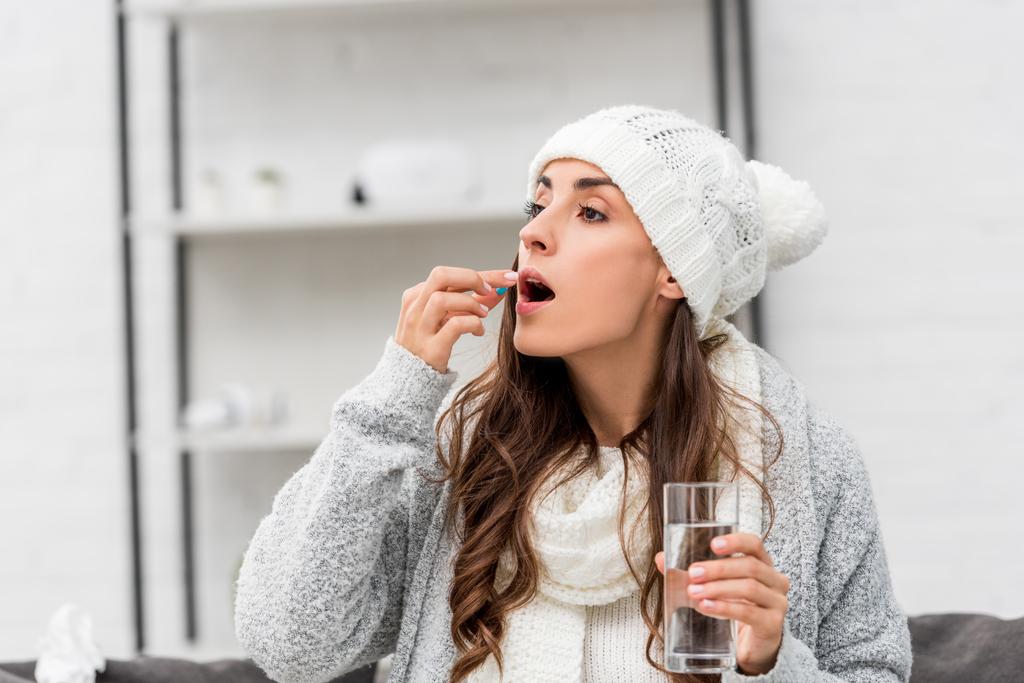 close-up portrait of sick young woman in warm clothes taking pill at home - Photo, Image