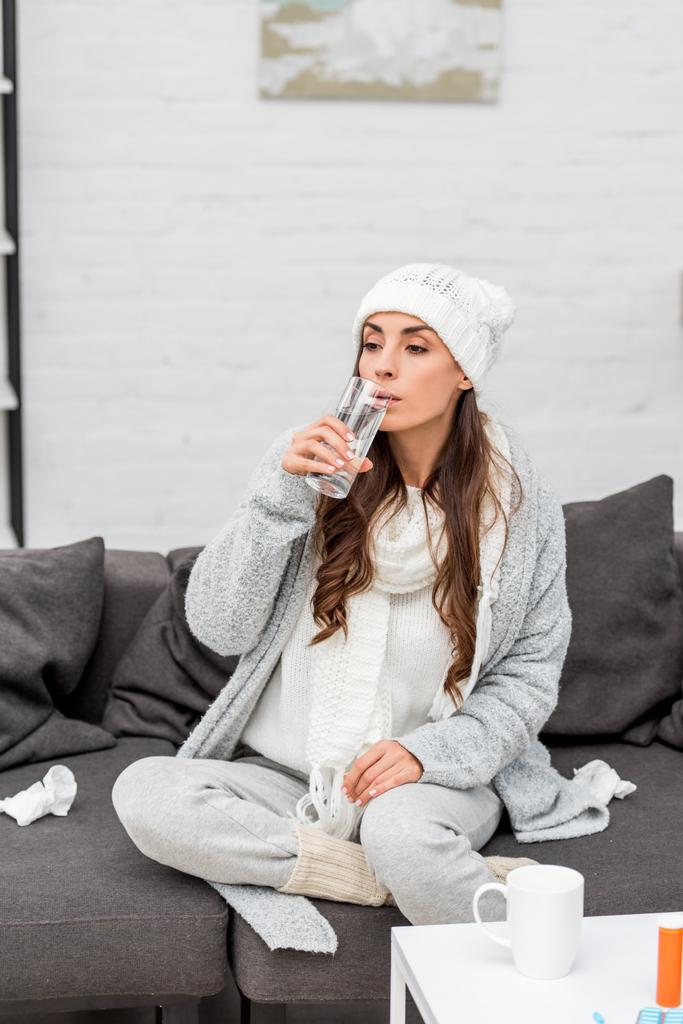 άρρωστος νεαρή γυναίκα σε ζεστά ρούχα πόσιμο νερό στον καναπέ στο σπίτι - Φωτογραφία, εικόνα