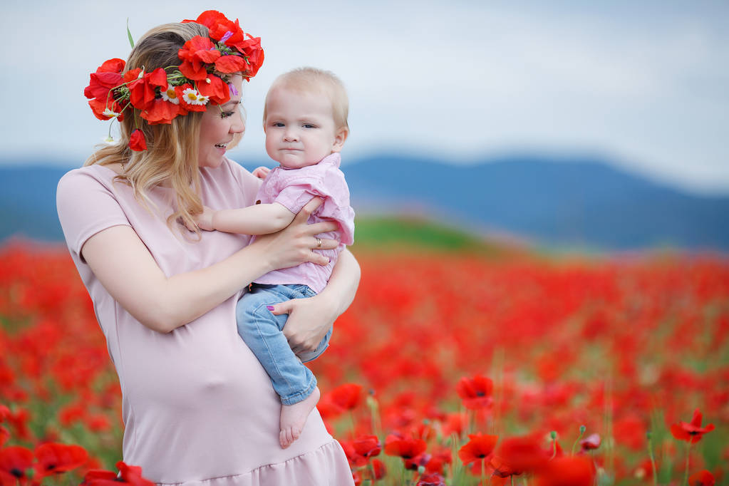 Feliz joven embarazada, rubia, vestida con un vestido rosa en la cabeza lleva una corona de flores frescas amapola pasa tiempo al aire libre en las montañas, en un vasto campo de amapolas rojas salvajes, a manos de su hijo pequeño
 - Foto, imagen