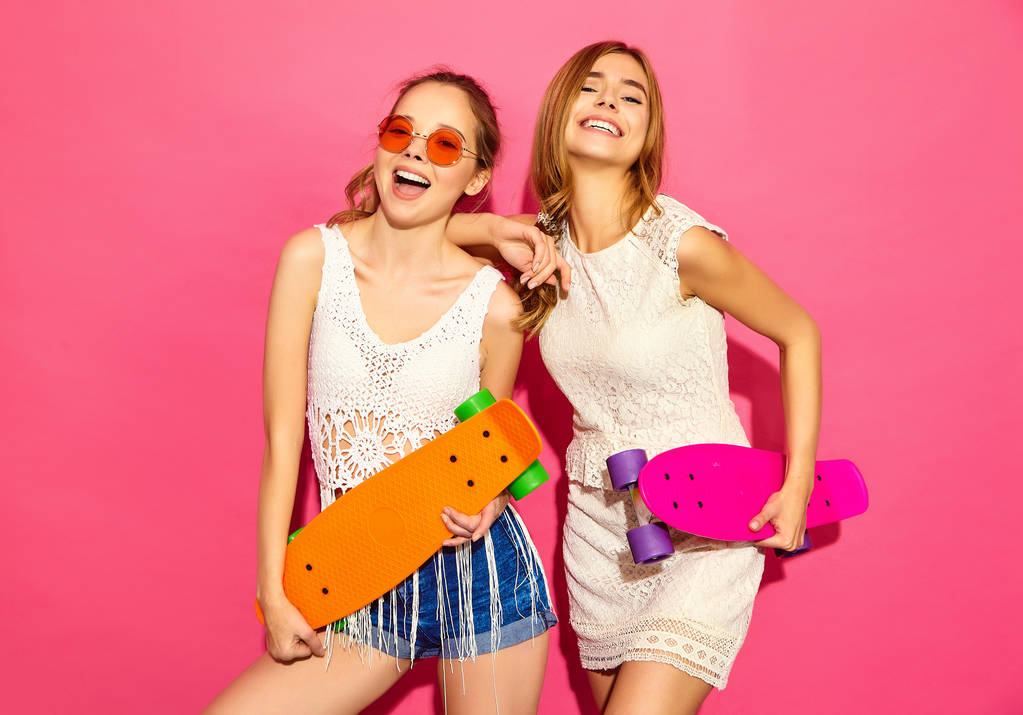 Дві молоді стильні усміхнені блондинки зі скейтбордами. Моделі в літньому хіпстерському білому одязі позують біля рожевої стіни в студії в сонцезахисних окулярах. Позитивна жінка
 - Фото, зображення