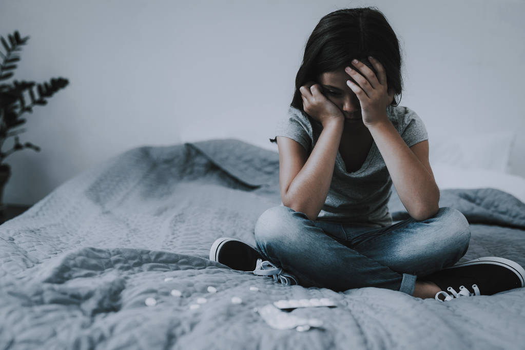 Little Girl Sits on Bed with Scattered Pills. Child on Gray Badcover with Crosssed Legs Near Opened Packing of Medicines Covers Face with Hands. Suicidal Tendencies and Unhappy Childhood Concept - Photo, Image